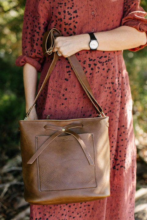 Wearing a rust-patterned dress, a person carries Urban Southern’s The Lexington Bow—a brown leather crossbody with a front pocket and bow detail—paired with a black wristwatch. Blurred green foliage forms the background.