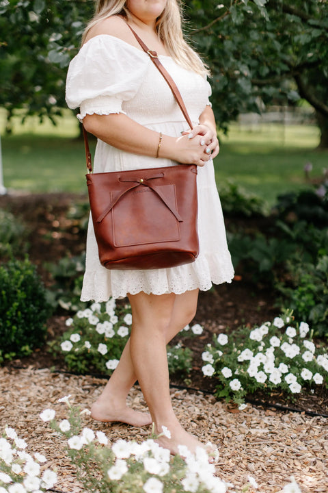 A woman in a white, off-the-shoulder dress stands barefoot among white flowers, holding Urban Southern’s The Lexington Bow crossbody. Her face is hidden, set against a lush green outdoor background.