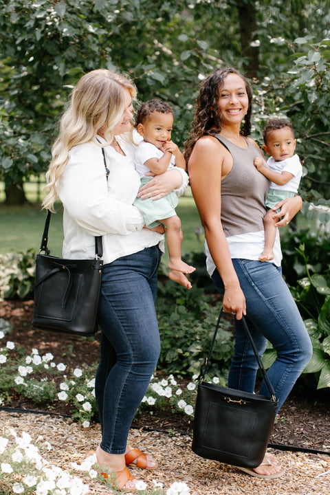 Two women stand in a garden, each holding a toddler. Both carry Urban Southern’s The Lexington Bow crossbody purse. The toddlers wear matching white shirts and light green shorts surrounded by flowers and greenery.