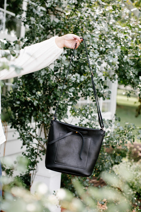 Someone holds up Urban Southerns The Lexington Bow, a black leather crossbody bag, by its strap outdoors with greenery and a white building blurred in the background.