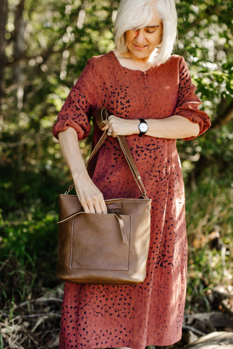 A woman with white hair in a red patterned dress stands outdoors, reaching into her Lexington Bow crossbody bag by Urban Southern, surrounded by trees and greenery.
