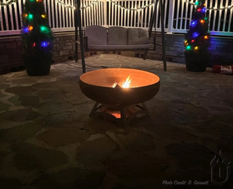 At night, a cozy fire glows in the Ohio Flame Liberty Fire Pit With Hollow Base (Made in USA) on a stone patio. Two potted trees with colorful string lights stand near a swing bench beneath softly glowing patio lights.