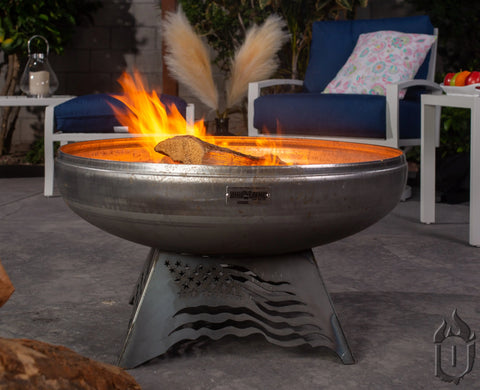 The Liberty Fire Pit Stars and Stripes *Limited Release* by Ohio Flame, with a striking American flag cutout stand, burns brightly on a patio surrounded by outdoor furniture and plants. Made in the USA.
