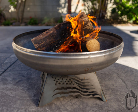 The Ohio Flame Liberty Fire Pit Stars and Stripes *Limited Release* (Made in USA) holds burning firewood, its flames glowing brightly on a concrete patio outdoors, standing out against nearby shrubs and a wall.