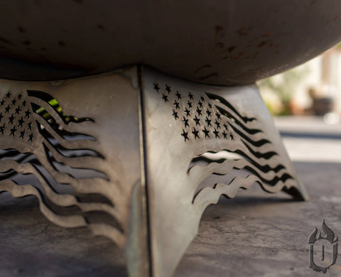 Close-up of the Ohio Flame Liberty Fire Pit Stars and Stripes *Limited Release* (Made in USA) stand, showcasing a cutout U.S. flag design with stars and wavy stripes, set on a stone surface outdoors.