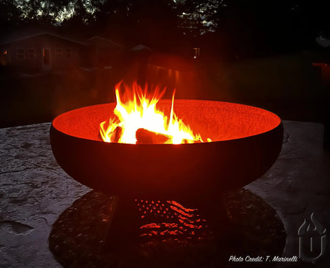 The Ohio Flame Liberty Fire Pit Stars and Stripes *Limited Release* (Made in USA) glows with bright orange flames on a patio at dusk, silhouetted houses and trees visible in the background. Photo credit: T. Marinelli.