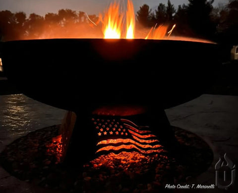A blazing fire glows in the Liberty Fire Pit Stars and Stripes *Limited Release* by Ohio Flame, its American flag base lighting up the evening outdoors with patriotic warmth beneath trees and a night sky. Made in USA.