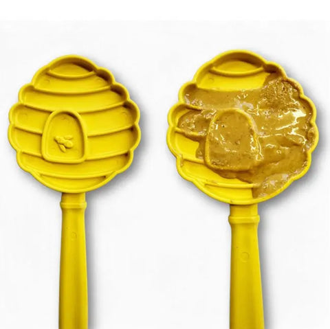 Two yellow SodaPup Lick Stick Hand-Held Interactive Enrichment Toys are shown—one clean, the other topped with peanut butter. Both feature bee hive shapes, handles, and raised bee designs for engaging food-based enrichment activities.