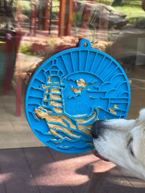 A dog enjoys peanut butter on the blue, circular SodaPup Lighthouse EMat Enrichment Lick Mat with suction cups, featuring a lighthouse and seagull design. This mat provides mental stimulation and attaches easily to glass doors.