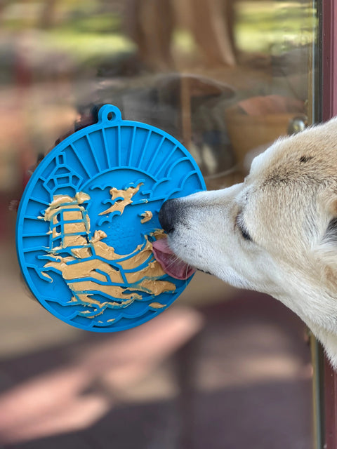 A light-colored dog enjoys a Dogs Brain Challenge, licking peanut butter from a blue, round Lighthouse EMat Enrichment Lick Mat With Suction Cups by SodaPup attached to a glass door; indoor and outdoor areas are visible in the blurred background.