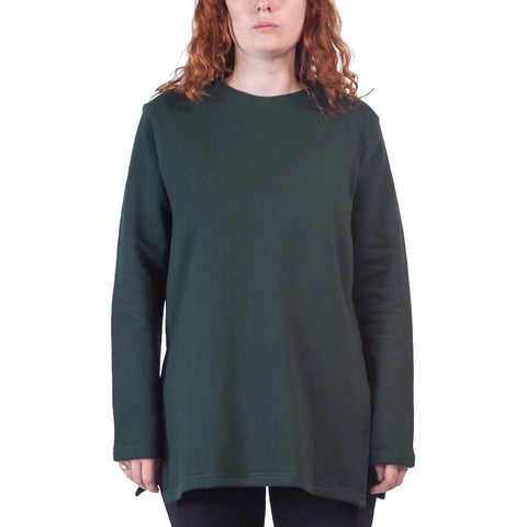 A person with wavy red hair wears the Flint Street Fleece Pullover by Spiritex | Sustainable Apparel in dark green organic cotton, paired with black pants, standing against a white background.