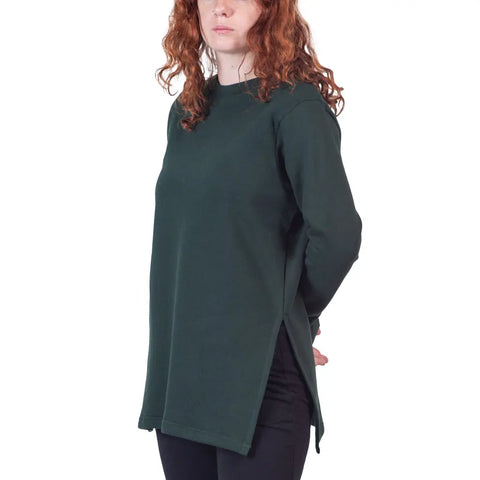A person with long curly red hair models the Flint Street Fleece Pullover by Spiritex | Sustainable Apparel, a dark green lightweight tunic with side slits, standing hands behind back against a plain white background.