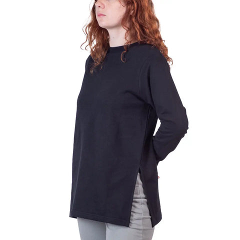 A person with long, curly red hair wears the Flint Street Fleece Pullover by Spiritex | Sustainable Apparel in black organic cotton fleece with side slits, paired with light gray pants, standing against a plain white background with hands behind their back.