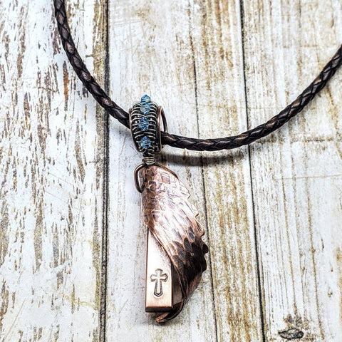 The Memorial Birthstone Angelwing Necklace-Limited Special Edition 2020-2024 by Alexa Martha Designs features a copper angel wing pendant with cross and blue wire, displayed on a braided dark leather cord.