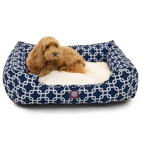 A small, fluffy brown dog rests on the Majestic Pet Links Contemporary Lounge Bed, featuring a cozy blue cover with a white geometric pattern and a soft premium fiberfill cushion inside.