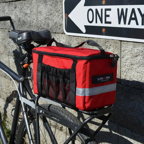A Lone Peak Basic Rack Pack from Lone Peak Packs is attached to a rear bike rack. The bike is parked near a stone wall, with part of a One Way street sign visible in the background.