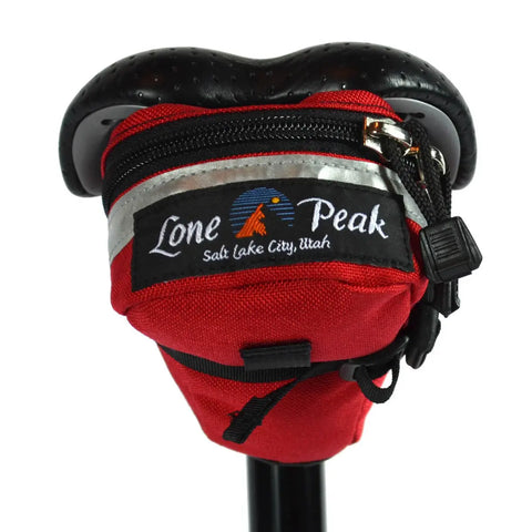 A red Lone Peak Packs Tool Pouch Seat Bag fits under a black bike seat, with a zipper, reflective strip, and Lone Peak logo with Salt Lake City, Utah—an essential accessory for carrying tools or gear.