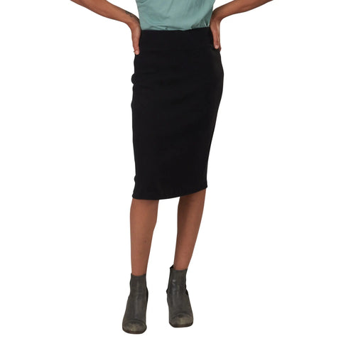 A person wears a light green top, the Long Andy Skirt in black from Spiritex | Sustainable Apparel, and gray ankle boots, standing hands on hips against a white background—an effortlessly chic look for modern office wear.