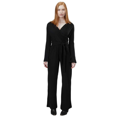 A woman with long red hair stands facing forward, wearing the Spiritex Long Sleeve Surplice Jumpsuit in Organic Cotton/Tencel with a waist tie and black shoes, set against a plain white background.