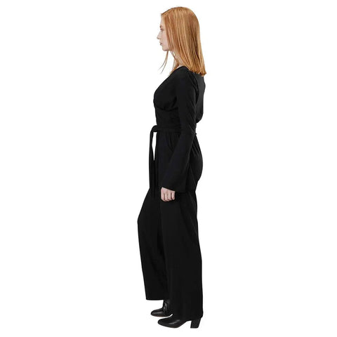 A woman with straight red hair stands in profile, wearing the Spiritex Long Sleeve Surplice Jumpsuit in Organic Cotton/Tencel with a belt and black heeled boots, against a plain white background.
