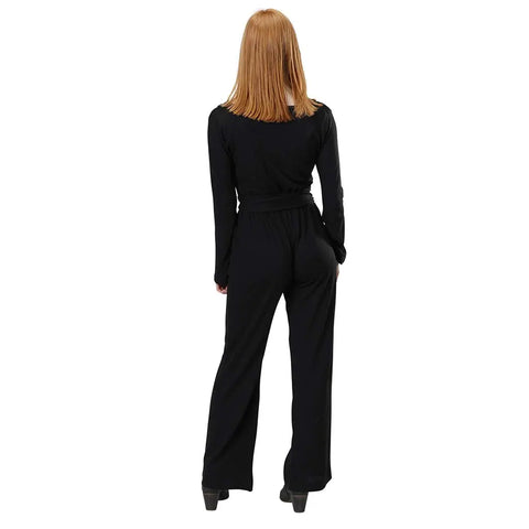 A person with straight, shoulder-length reddish hair faces away, modeling the Spiritex Long Sleeve Surplice Jumpsuit in Organic Cotton/Tencel with black heels against a plain white background.