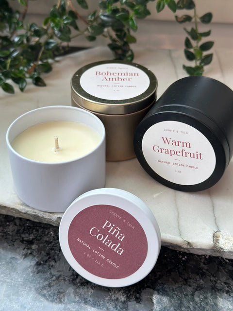 Three Shanti + Tula Lotion Candles with Shea, Cocoa & Mango Butter—clean-scented Bohemian Amber, Warm Grapefruit, and Piña Colada—are displayed on marble with greenery, offering a USA-made self-care massage oil and skin hydrator.
