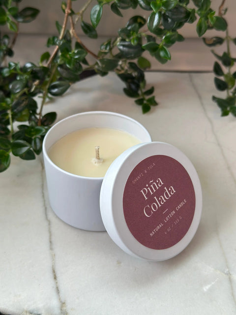 The Shanti + Tula Lotion Candle with Shea, Cocoa & Mango Butter sits open on a marble surface with leafy vines behind it—a USA-made, clean-scented candle perfect for massage oil and skin hydration during self-care.