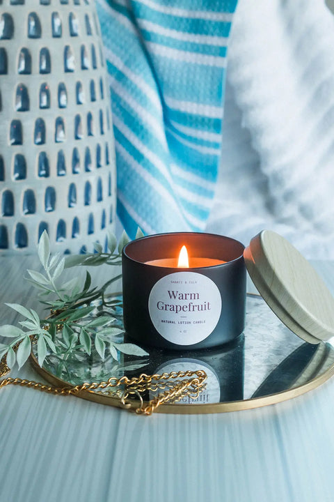 A lit Shanti + Tula Lotion Candle with Shea, Cocoa & Mango Butter sits on a round mirror with its wooden lid nearby, beside a gold chain and green leaves. Blue and white fabrics in the background evoke a relaxing self-care experience.