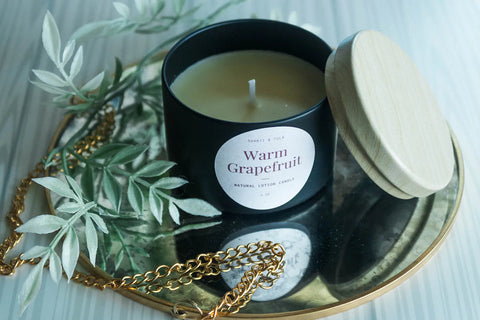 A black Shanti + Tula Lotion Candle with Shea, Cocoa & Mango Butter rests on a gold tray with its wooden lid, surrounded by leaves and a gold chain, creating a self-care vibe. Enjoy clean scents and skin hydration with this USA-made massage oil candle.