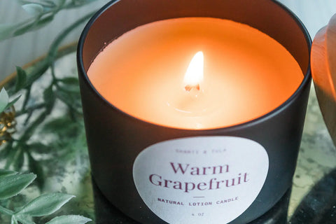 A lit Shanti + Tula Lotion Candle with Shea, Cocoa & Mango Butter rests on a reflective surface near green leaves—this USA-made, clean-scented candle melts into massage oil for skin hydration and relaxing self-care.