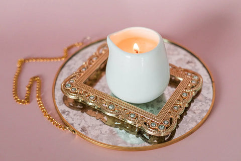 The Shanti + Tula Lotion Candle with Shea, Cocoa & Mango Butter sits in a white ceramic holder on a gold-framed mirror atop a pink surface—perfect for soothing self-care, massage oil, and skin hydration.