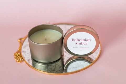 A Shanti + Tula Lotion Candle with Shea, Cocoa & Mango Butter rests open on a gold-edged mirrored tray against a soft pink background—a USA-made, clean-scented massage oil candle perfect for elevating your self-care routine.