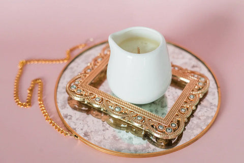 A Shanti + Tula Lotion Candle with Shea, Cocoa & Mango Butter sits on a gold frame atop a marble and gold tray with chain handle, creating a soothing self-care vibe against a light pink background.
