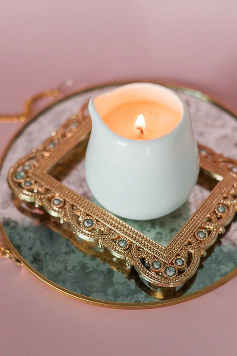 The Shanti + Tula Lotion Candle with Shea, Cocoa & Mango Butter rests in a white holder on a jeweled gold frame atop a round, gold-rimmed mirror—an elegant USA-made massage oil candle to elevate your self-care routine.