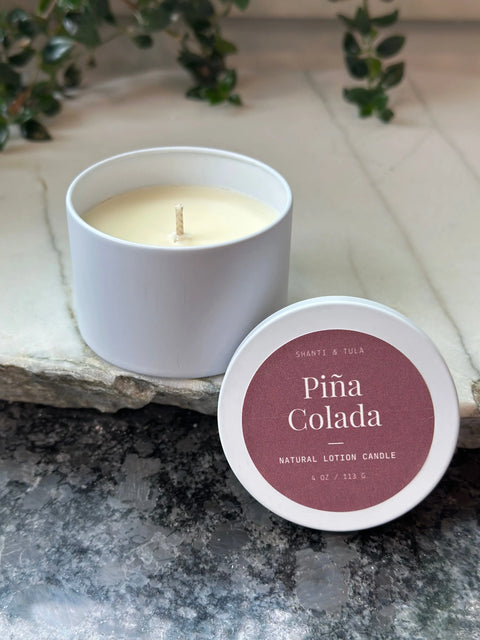 A white Shanti + Tula Lotion Candle with Shea, Cocoa & Mango Butter sits in a round container on marble. The lid, nearby, displays a maroon label: “Piña Colada Natural Lotion Candle, 4 oz / 113 g.” Green leaves are in the background.