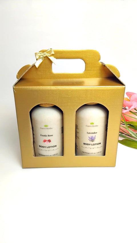 A gold gift box with a handle and bow displays two bottles of Forev'Herbs Lotion Gift Set, labeled Exotic Rose and Lavender. Pink flowers are placed beside the set on a white background.