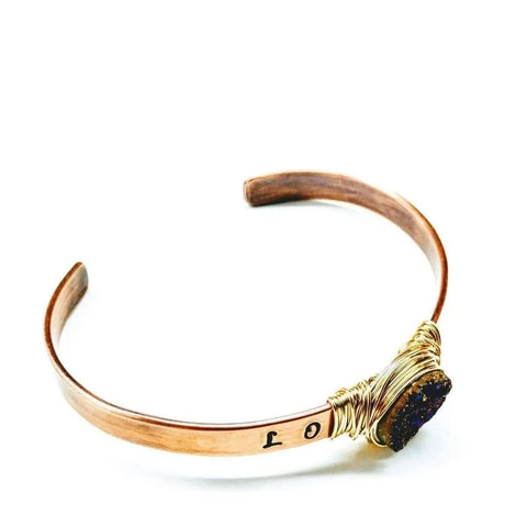 The LOVE Stamped Gold Wire Wrapped Purple Druzy Copper Cuff by Alexa Martha Designs is a handmade bracelet featuring gold wire wrapping around a dark, textured stone. “0 1” is stamped inside and it’s showcased on a plain white background.