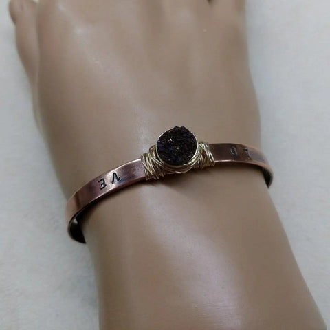 The LOVE Stamped Gold Wire Wrapped Purple Druzy Copper Cuff by Alexa Martha Designs features engraved letters, a textured purple druzy agate wrapped in gold-filled wire at the center, and is shown worn on a mannequin’s wrist.