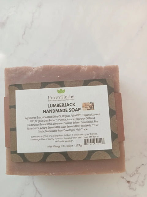 A bar of Lumberjack Handmade Soap by Forev'Herbs, a masculine exfoliating soap, sits on a white surface. The label lists ingredients and notes that this fair trade soap is made with sustainable practices.