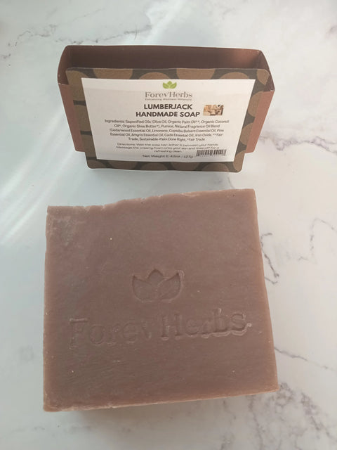 A square brown bar of Forev'Herbs Lumberjack Handmade Soap, embossed with the brand name, sits on a white surface. Behind it is a label listing the soap’s natural ingredients.