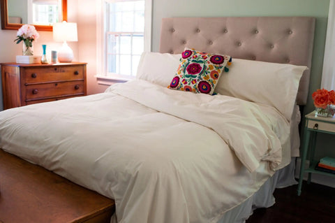 A neatly made bed features the Cotton Duvet Cover Set by American Blossom Linens, paired with a floral pillow and tufted headboard. Nearby are a wooden dresser with flowers, a mirror, and a side table, all bathed in sunlight from the window.