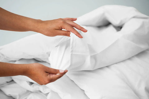 A person folds a white Cotton Duvet Cover Set by American Blossom Linens, with layers of bed linens in the background, showcasing the brands luxurious comfort and attention to detail.
