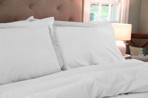 A neatly made bed featuring white pillows and the Cotton Duvet Cover Set by American Blossom Linens is set against a tufted headboard, with a window and lit table lamp visible in the softly lit room.