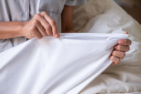 A person, visible only by their hands and torso, zips a pillowcase while sitting on cream bedding, highlighting the quality of the Cotton Duvet Cover Set by American Blossom Linens.