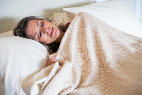 A woman with gray hair sleeps peacefully on her side, wrapped in the American Blossom Linens Herringbone Weave Cotton Blanket, resting her head on a white pillow.