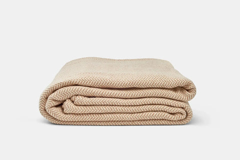The Herringbone Weave Cotton Blanket by American Blossom Linens features a subtle beige herringbone pattern, neatly folded against a white background—a cozy, inviting layer ideal for any space.