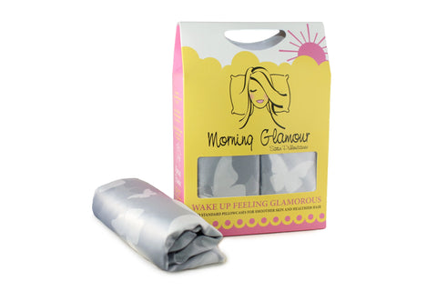 A box of Morning Glamour Satin Pillowcase Standard/Queen 2PK (Multiple Print Options) is shown next to a folded silver butterfly-print pillowcase. The yellow and pink packaging features a womans face and rising sun—great for reducing frizz overnight.