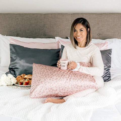 A woman in striped pajamas sits on a bed with Morning Glamour Satin Pillowcase Standard/Queen 2PK in gray and pink, holding a mug. She smiles, wrapped in a leopard print blanket, with a breakfast tray of croissants and strawberries beside her.