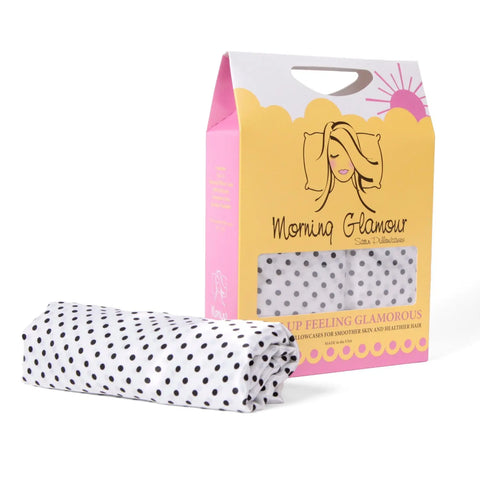 A folded white satin pillowcase with black polka dots sits in front of pink and yellow packaging with a woman’s face and Morning Glamour branding—this Standard/Queen 2PK helps reduce frizz for smoother hair while you sleep.