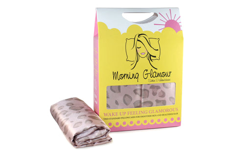 A box of Morning Glamour Satin Pillowcase Standard/Queen 2PK (Multiple Print Options) in pastel yellow and pink packaging features a woman’s face and sun. A folded silky leopard print pillowcase is in front, designed to reduce frizz.